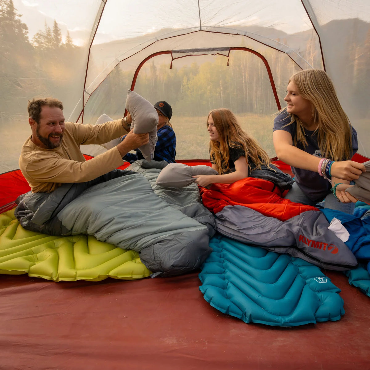 A family sits inside a tent having a pillow fight, with Klymit sleeping pads and sleeping bags spread across the floor.