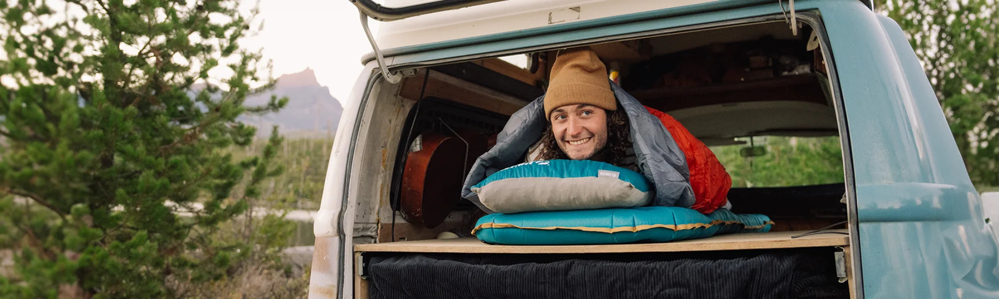 Smiling man wrapped in a sleeping bag lying on Klymit camping gear in the back of a vintage camper van at a mountain campsite.