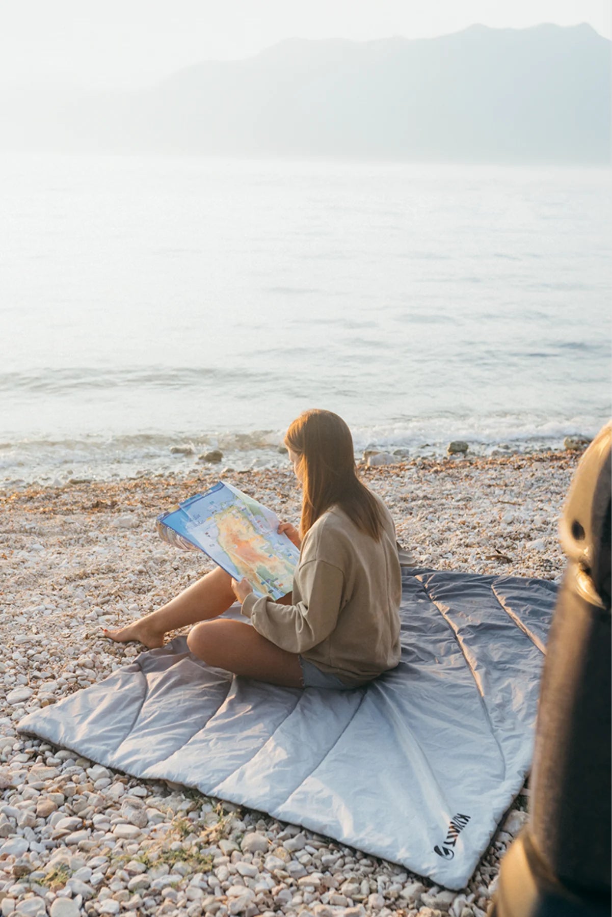 Woman reads a map while sitting cross-legged on a gray Klymit Horizon Overland Blanket spread out on a pebble beach at the water's edge.