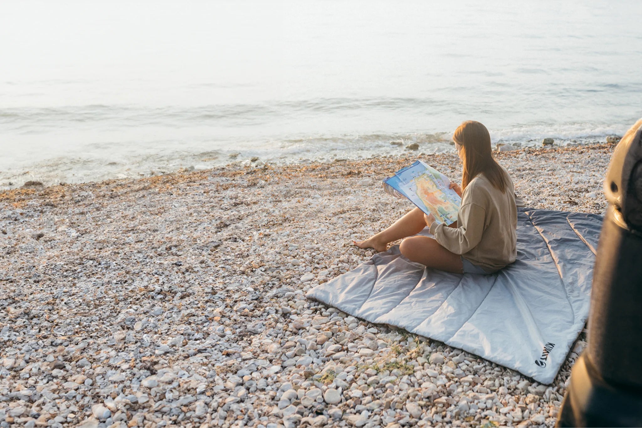 Woman reads a map while sitting cross-legged on a gray Klymit Horizon Overland Blanket spread out on a pebble beach at the water's edge.