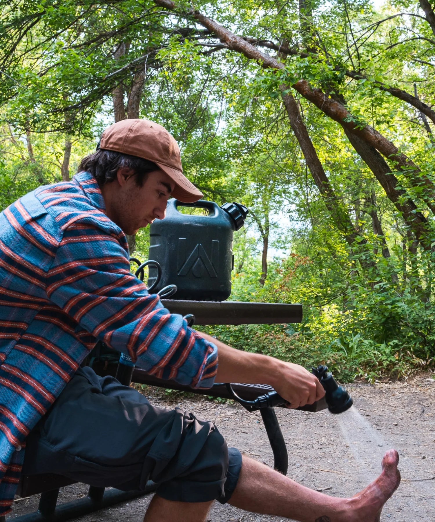 Man rinsing his foot using a WaterPORT GoSpout portable water tank with spray nozzle at a wooded campsite.